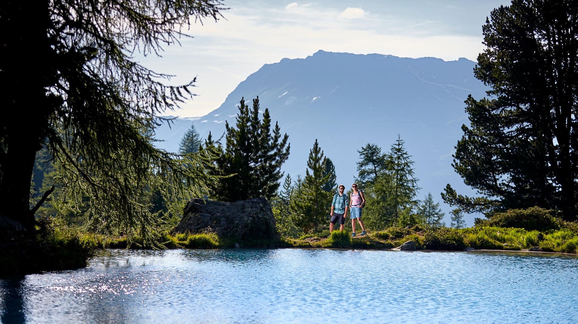 Bergsee mit klarem Wasser, umgeben von Bäumen, mit Blick auf Bergrücken – ruhige Naturkulisse in den Alpen.