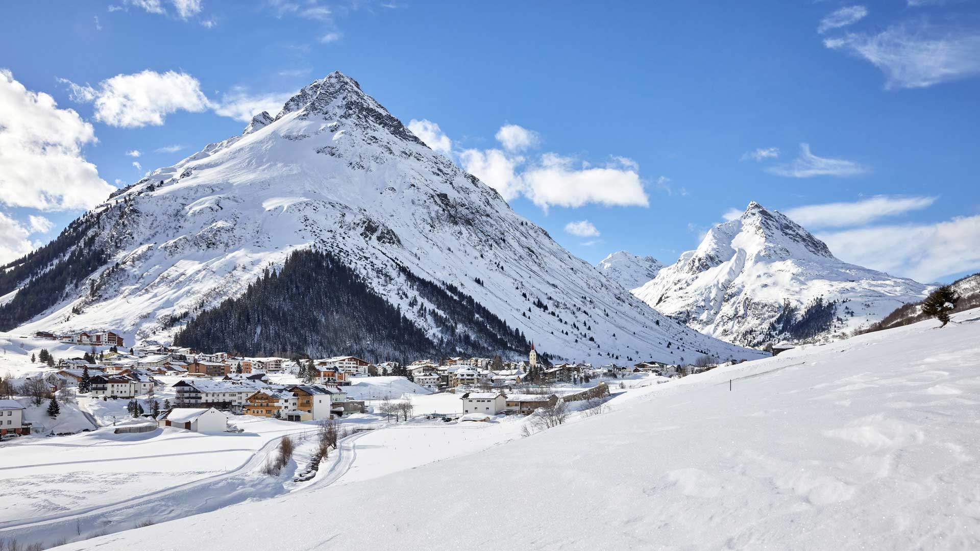 Verschneite Alpenlandschaft mit markanten Gipfeln, im Tal liegt ein malerisches Dorf unter blauem Himmel mit Wolken.