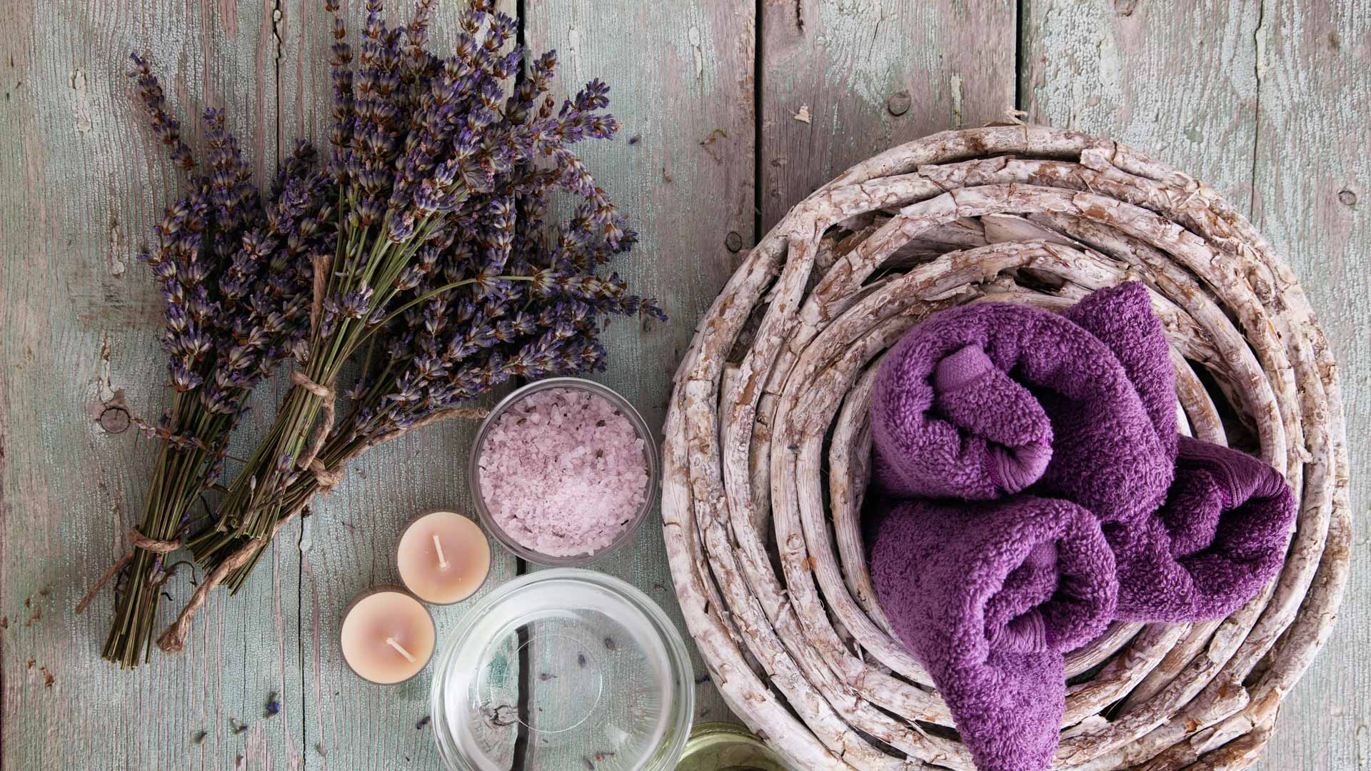 Wellness-Accessoires auf Holz: Lavendel, Kerzen, Handtuch im Korb und Badezusätze – natürliche und entspannende Szene.