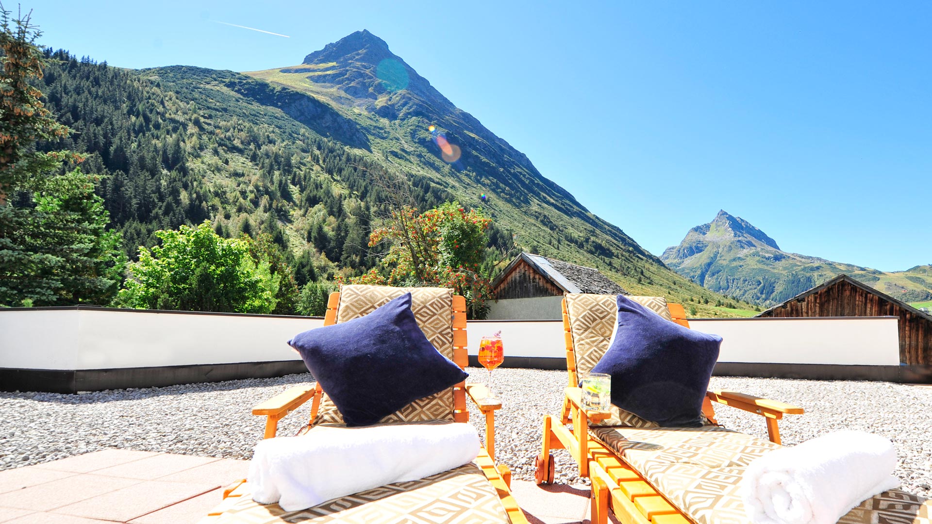 Zwei Sonnenliegen mit Kissen und Cocktails auf Terrasse mit Blick auf grüne Berge – Sommeridylle pur.