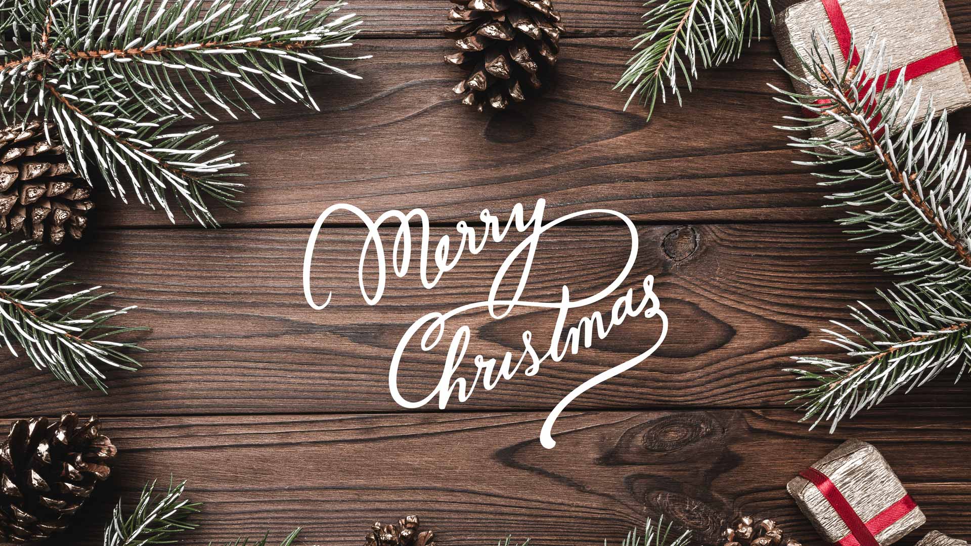 Weihnachtliche Dekoration mit Tannenzweigen, Zapfen, Geschenk und „Merry Christmas“-Schrift auf rustikalem Holzhintergrund.