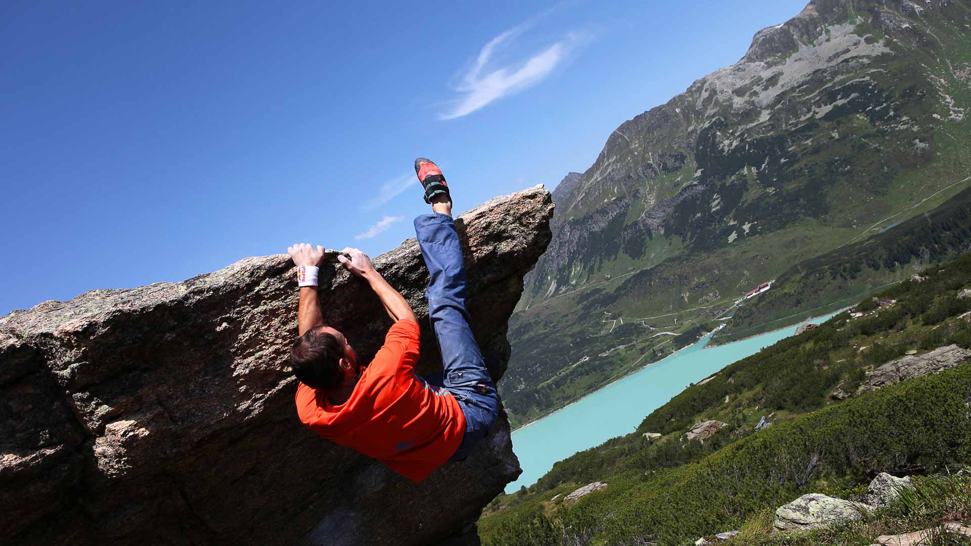 Boulderer auf einem Felsen mit blauer Hose und rotem Shirt, sonnige Berglandschaft.