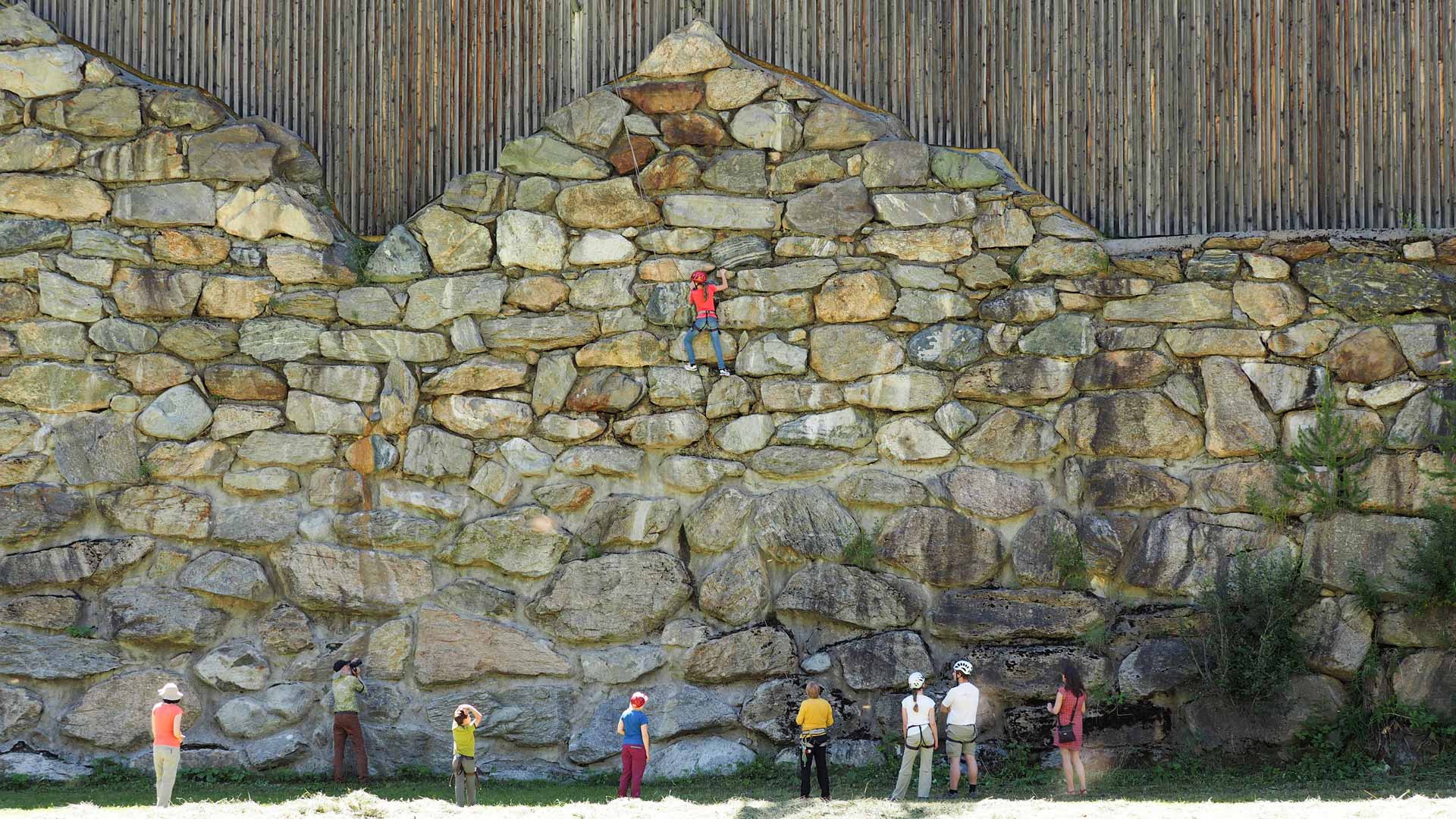 7 Kletterer stehen vor einer Felswand, ein Kletterer klettert an der Wand nach oben.